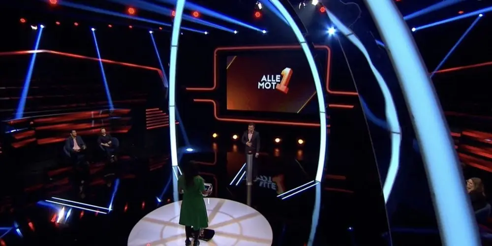 The image shows a brightly lit game show set with a glowing arch, a large screen displaying 